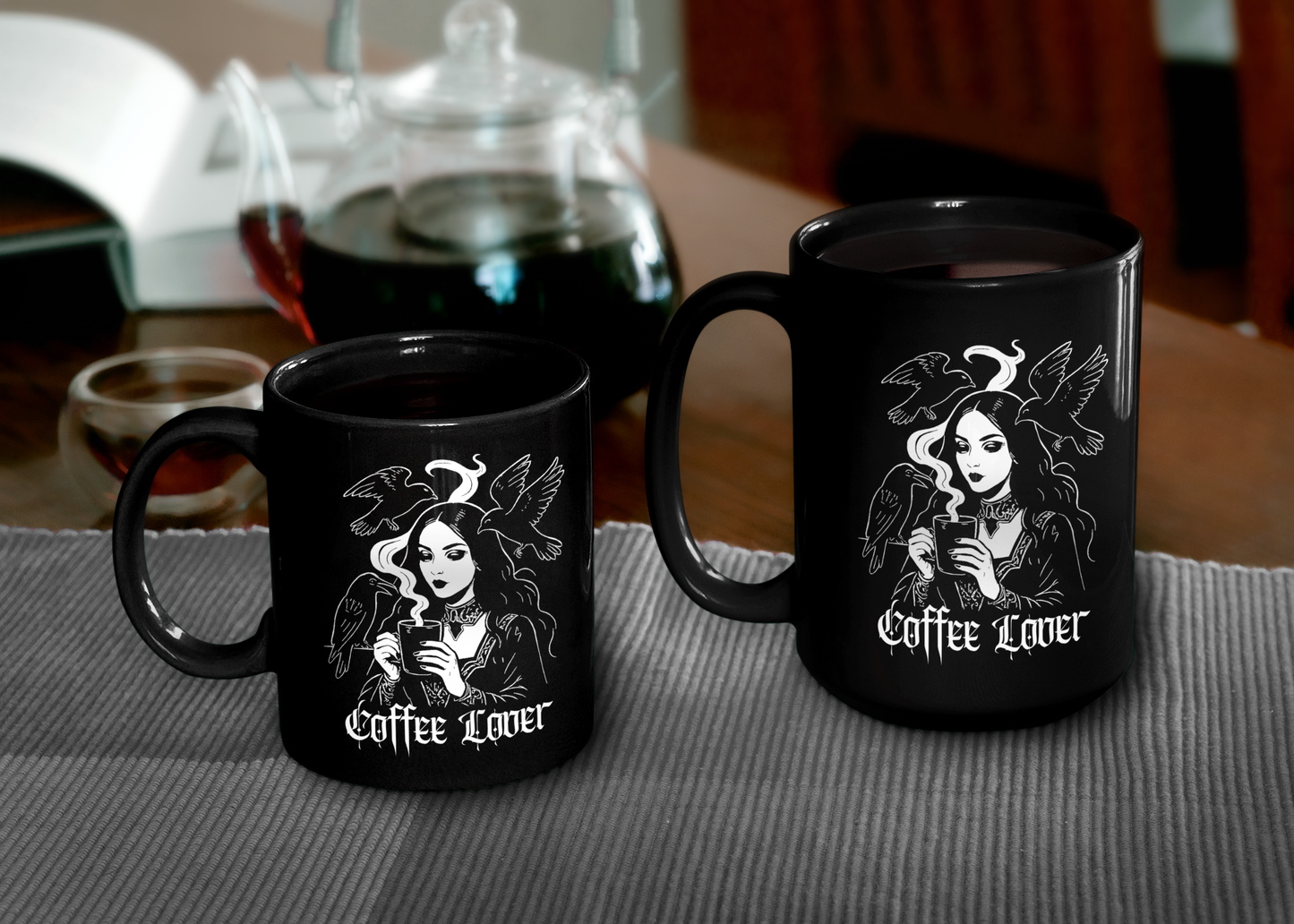 Two different sized black mugs with gothic 'Coffee Lover' design on a table with a teapot in the background.